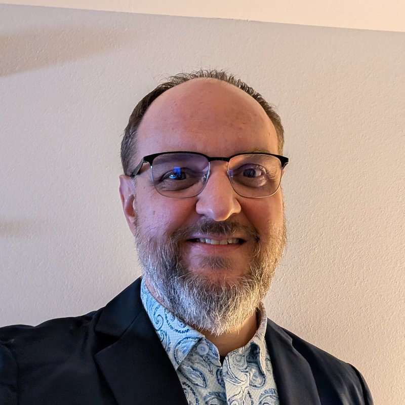 Kevin Wilk, smiling in glasses, a dark blazer, and a patterned shirt